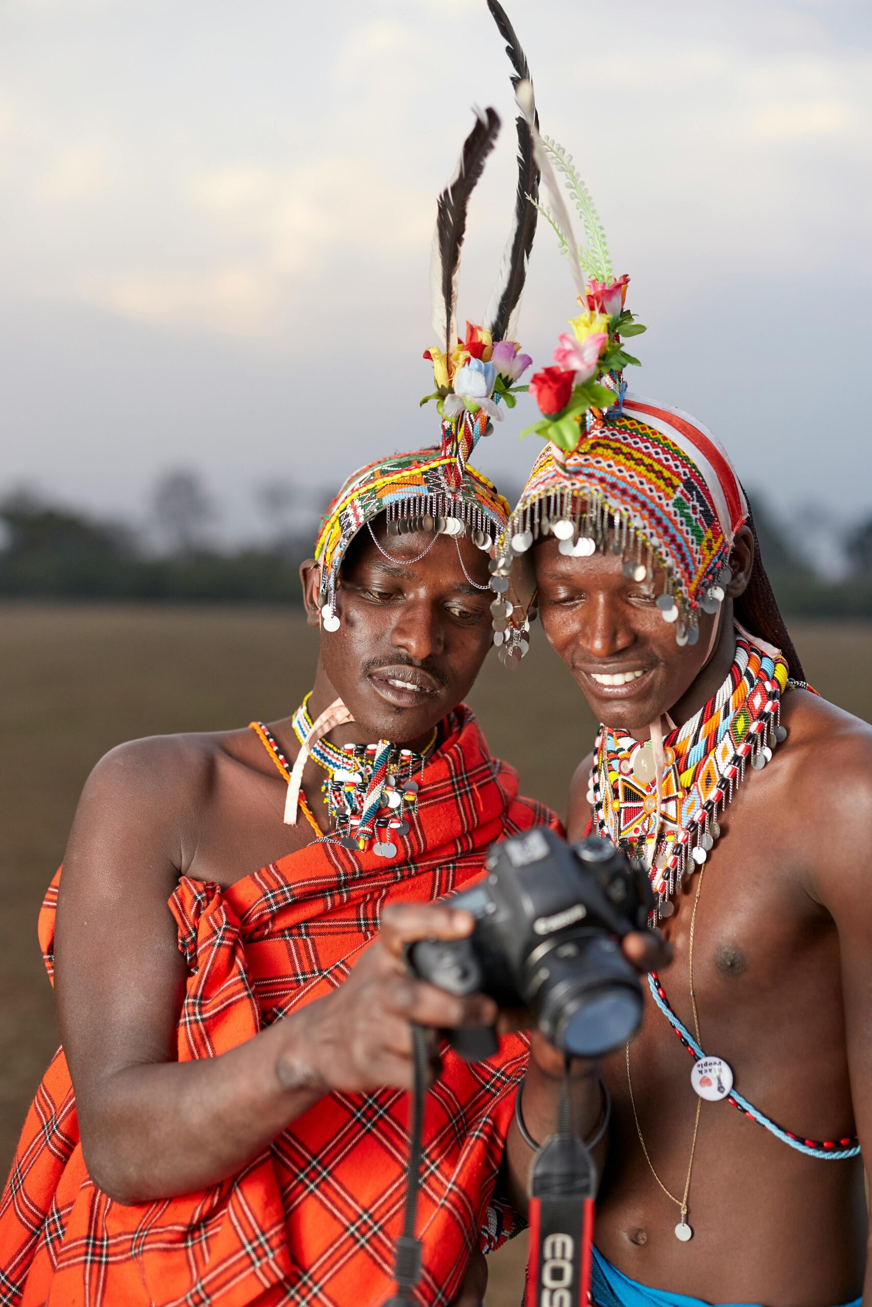 Two Masai men in vibrant traditional clothing examine a camera outdoors, showcasing culture.