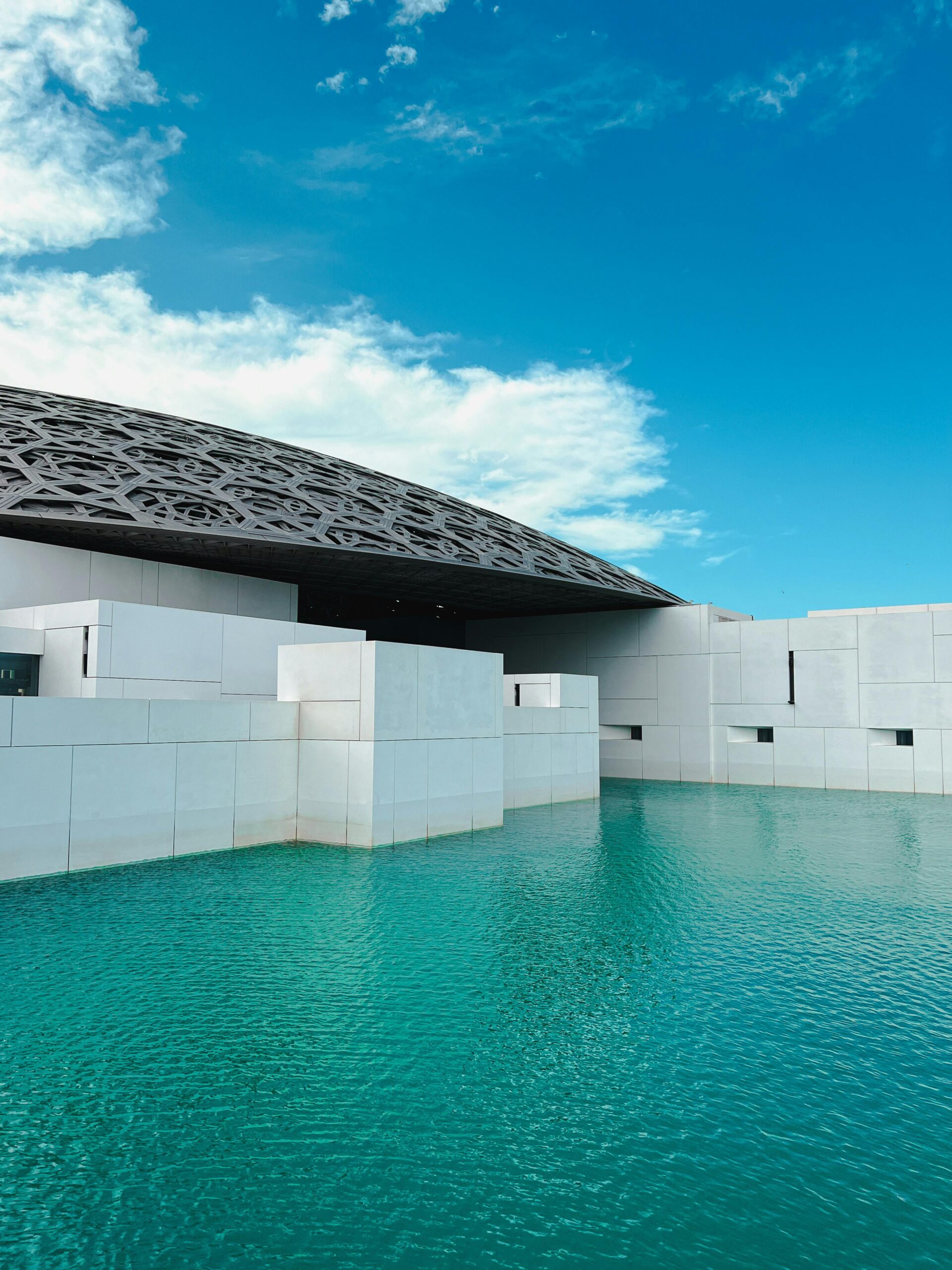 Explore the stunning modern architectural design of Louvre Abu Dhabi surrounded by serene water under a bright blue sky.
