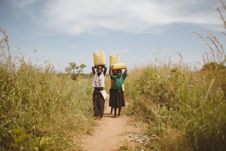 Two children walking through a rural pathway carrying water containers on their heads in Kitgum, Uganda.