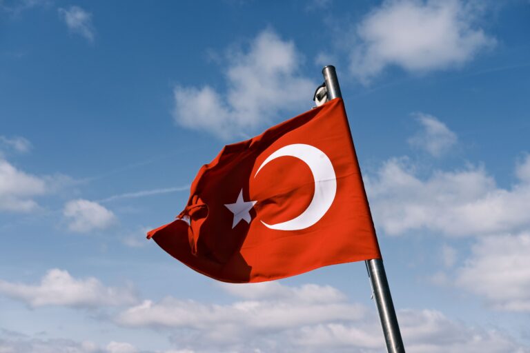 A vibrant Turkish flag waving vigorously against a bright blue sky with scattered clouds.